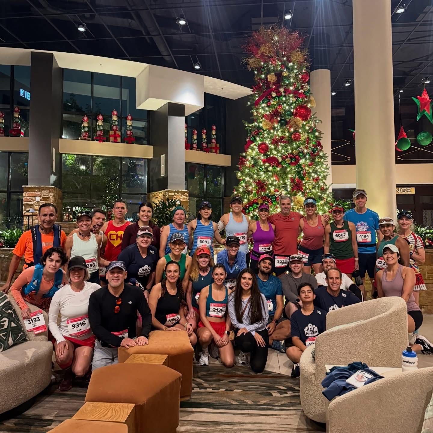 Large group of runners posing for a group photo indoors beside a tall Christmas tree with lights and ornaments.